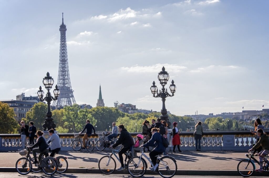 Cyclists and pedestrians share the Pont Alexandre III in Paris with the iconic Eiffel Tower rising in the distance under a partly cloudy sky, as ornate street lamps add to the city's historic charm.