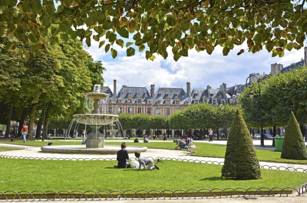 Place des Vosges in Paris on a sunny day, framed by leafy branches, features visitors relaxing near a classic fountain, with the distinctive red brick facades and steep slate roofs of the surrounding buildings.