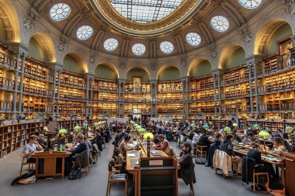 The grandiose reading room of the Bibliothèque nationale de France, filled with patrons at wooden desks under green reading lamps, surrounded by towering bookshelves and ornate architecture with a glass dome ceiling.