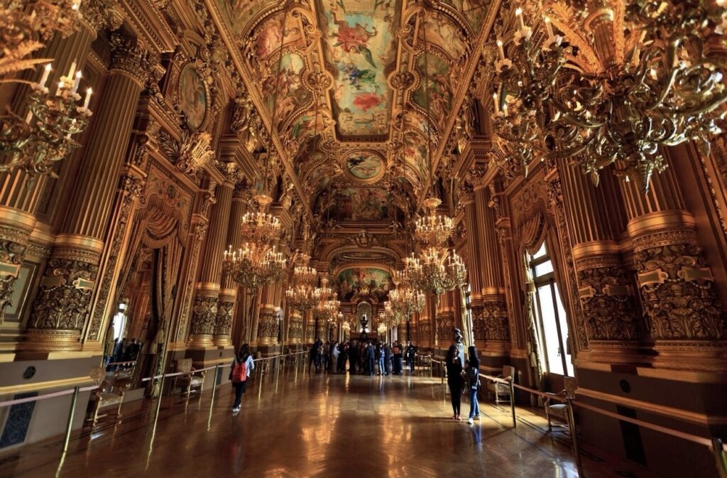 Visitors exploring the ornate Grand Foyer of the Palais Garnier, Paris, with its opulent golden columns, intricate frescoes, and glittering chandeliers, reflecting the grandeur of classic French architecture.