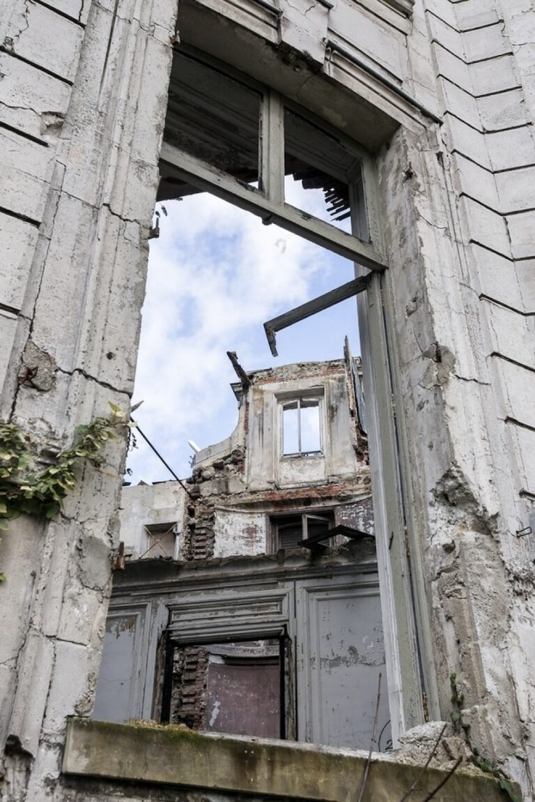 View through a broken window frame of a dilapidated building, showcasing the sky, with the remnants of a decaying structure and exposed bricks in the foreground.