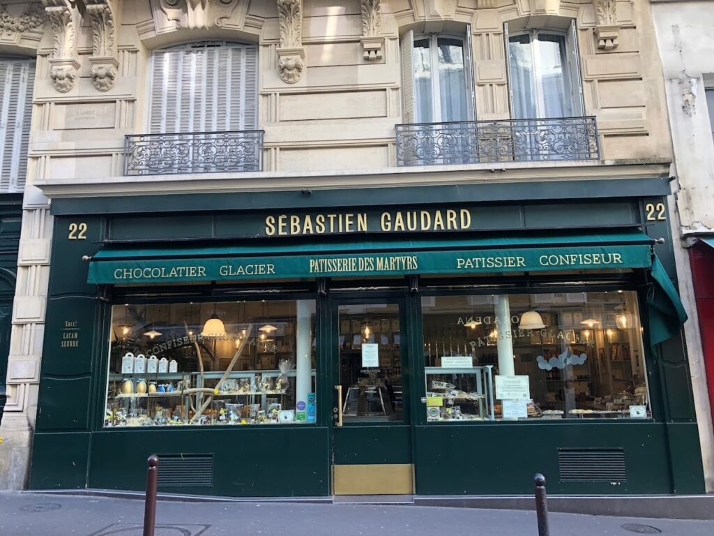 hidden gems in paris: The storefront of Sebastien Gaudard patisserie in Paris, featuring a green awning with gold lettering, and a display window full of desserts. Above the shop is a traditional Parisian building facade with intricate sculptures and a wrought-iron balcony.