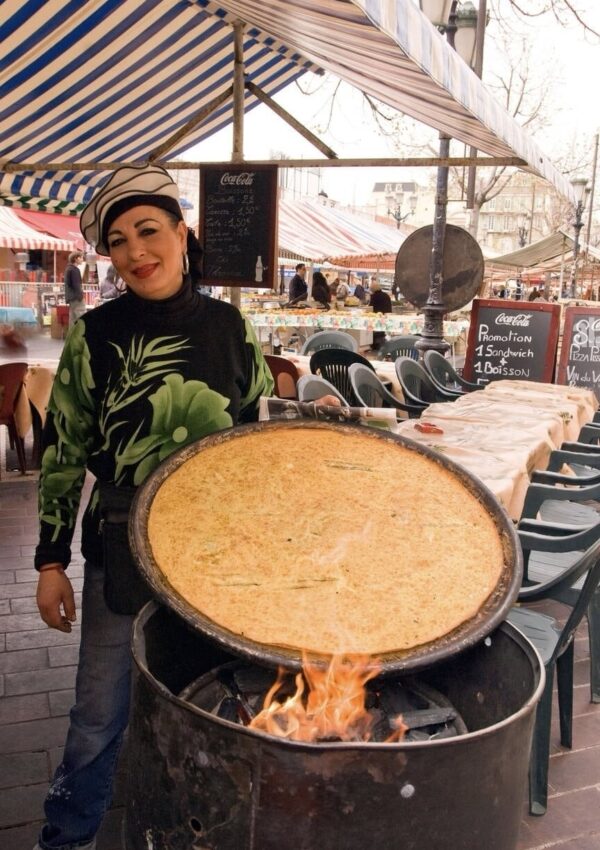 A smiling vendor stands beside a large, round griddle with a freshly cooked socca over an open flame at a market stall in Nice. Behind her, the market bustles with activity under striped awnings, with a blackboard advertising food and drink specials.