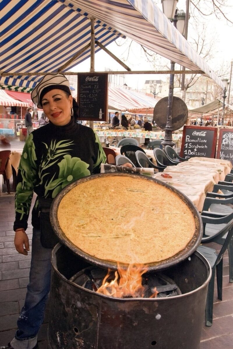 A smiling vendor stands beside a large, round griddle with a freshly cooked socca over an open flame at a market stall in Nice. Behind her, the market bustles with activity under striped awnings, with a blackboard advertising food and drink specials.
