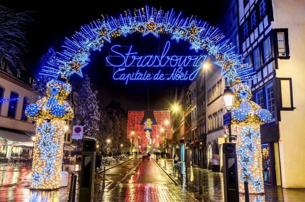 Festive holiday decorations light up a wet street in Strasbourg at night, with a radiant arch reading 'Strasbourg Capitale de Noël.' The scene is embellished with blue and gold illuminated stars and Christmas figures, reflecting off the glistening pavement alongside traditional half-timbered houses.