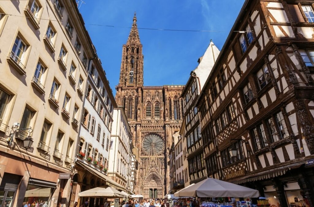 A bustling street in Strasbourg leads to the imposing Strasbourg Cathedral, which towers above with its intricate gothic façade and towering spire against a clear blue sky. Flanked by traditional European buildings and half-timbered houses, the scene captures the lively urban atmosphere juxtaposed with historic architecture.