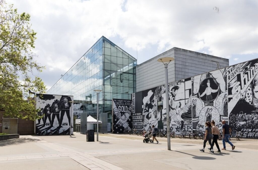 The Musée d'Art Moderne et Contemporain de Strasbourg (MAMCS) with its modern glass façade reflecting the sky. In the foreground, a striking black and white mural adds a dramatic touch to the urban landscape, while visitors stroll by, enjoying the blend of contemporary art and architecture.