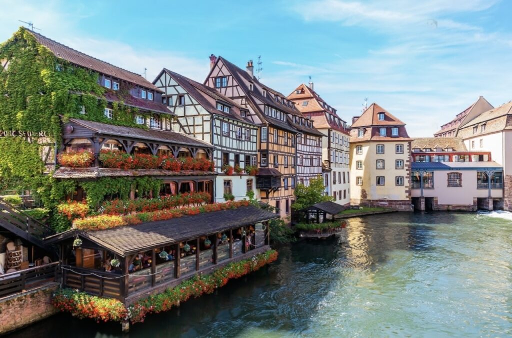 The quaint and charming quarter of Petite France in Strasbourg, with half-timbered houses draped in green ivy and red flowers. The buildings are reflected in the gently flowing river, and a covered restaurant terrace sits invitingly on the water's edge, capturing the essence of this picturesque historic area.