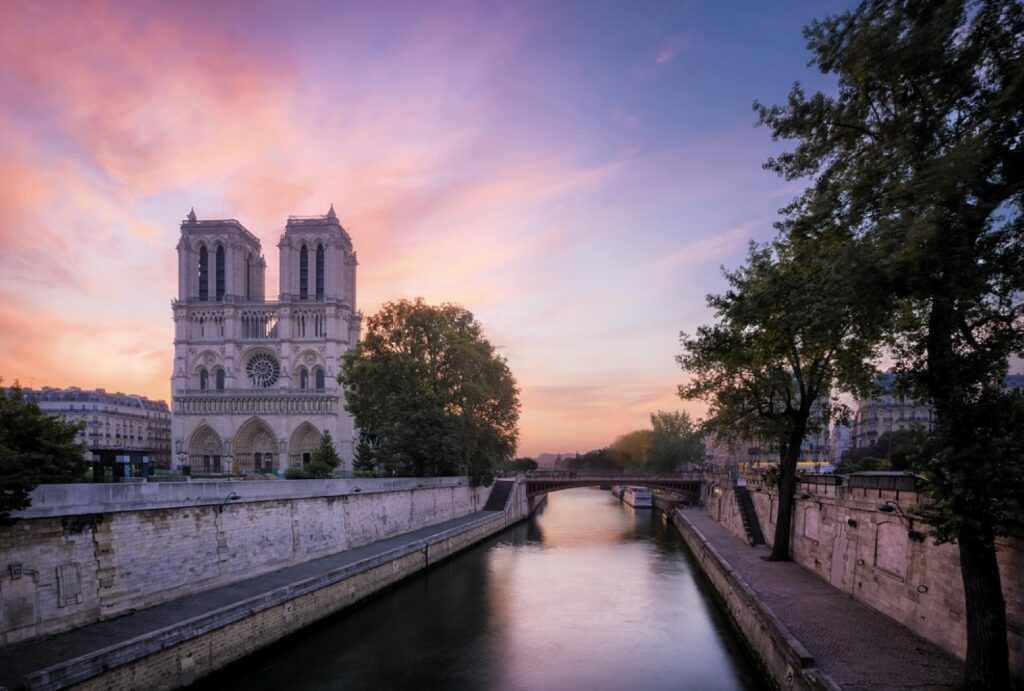 Sunrise at Notre Dame Cathedral with the sky painted in shades of pink and purple, reflecting over the calm waters of the Seine River, framed by lush trees and the stone architecture of the riverbank.