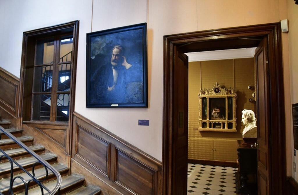 Inside the Victor Hugo House in Paris, a painting of the illustrious writer adorns the wall beside a wooden staircase, leading to a room displaying a classical bust and ornate religious artifacts.
