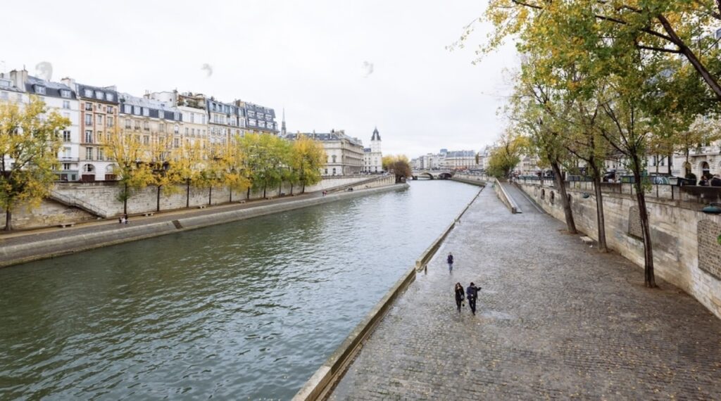 A peaceful scene along the River Seine in Paris with a few people walking on the cobblestone quay, flanked by trees with early autumn leaves and historic Parisian buildings, under a cloudy sky.