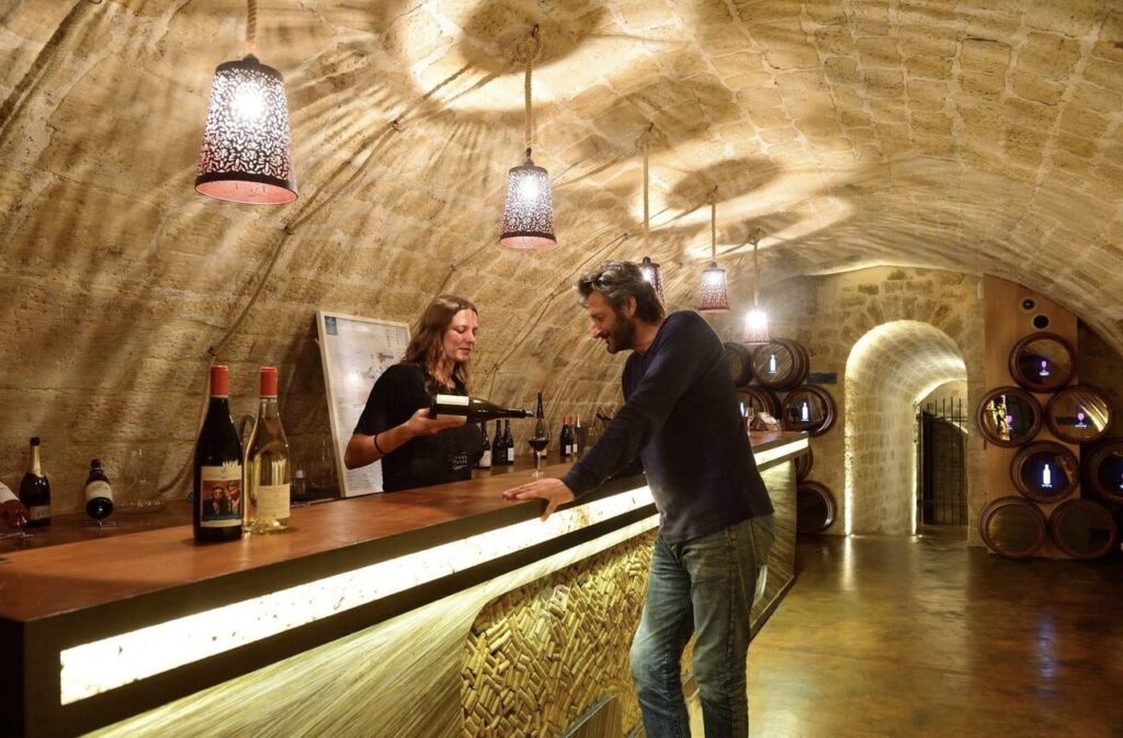 A cozy wine cellar tasting room with vaulted stone ceilings, a female server presenting a bottle to a male customer at the bar, surrounded by wine barrels and bottles on display under warm pendant lighting.