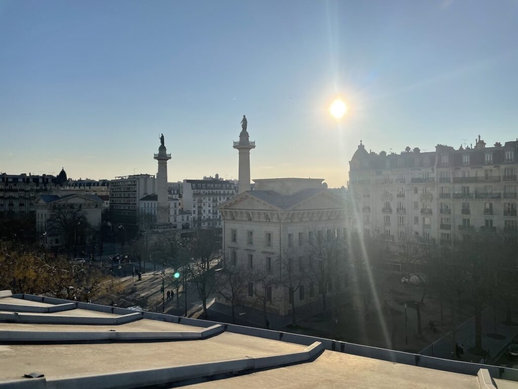 airbnbs in paris: Sunrise view over a serene cityscape, showcasing the soft silhouette of classical European architecture with distinct rooftops and two prominent statues atop pillars. The sun casts a warm glow and long shadows, offering a peaceful start to the day in the city.