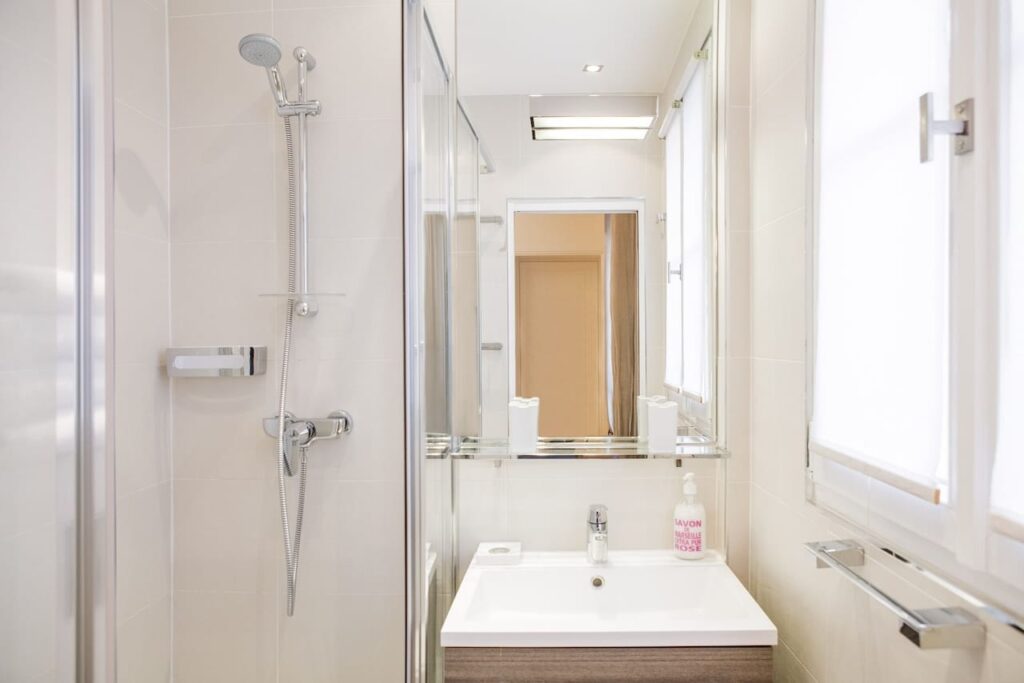 airbnbs in paris: Modern and bright bathroom interior with clean white tiles, featuring a glass shower enclosure with a mounted adjustable shower head, a rectangular sink with a large mirror above it reflecting the room's entrance, and amenities including a bottle of 'Savon Marseille Rose' hand soap.