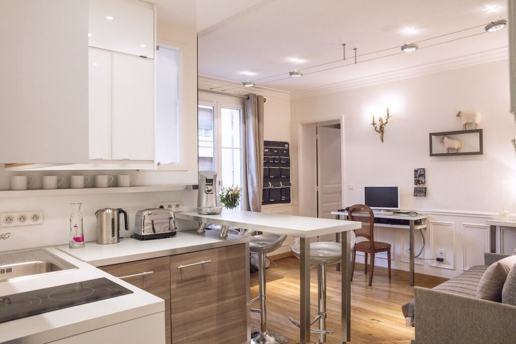 airbnbs in paris: Elegant open-plan kitchen and living space with classic wood flooring, featuring white cabinetry and countertops, modern appliances including a kettle and toaster, and a breakfast bar with high stools. There's a cozy work corner with a wooden desk and chair, and wall art depicting sheep adds a touch of whimsy to the room.
