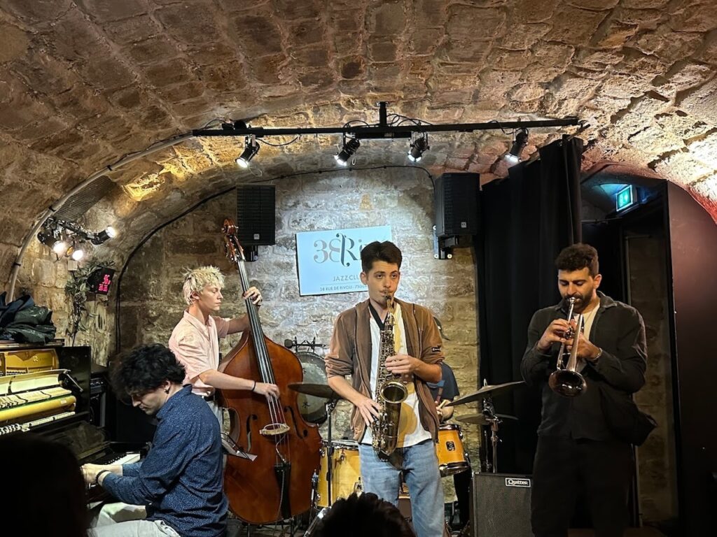 A live jazz quartet performs in the cozy ambiance of 38Riv Jazz Club, with exposed stone arches overhead. The ensemble features a double bassist, a pianist, a saxophonist, and a trumpeter deeply engaged in their music.