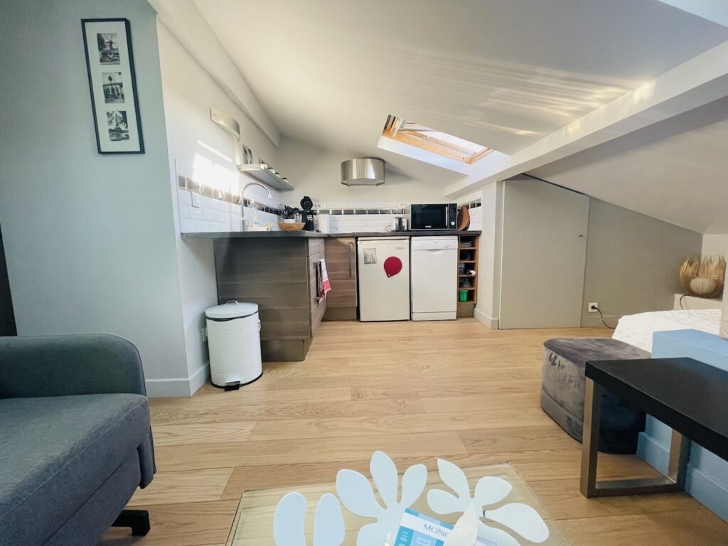 airbnbs in paris: Chic and compact studio apartment with a sloped ceiling and skylight, showcasing a tidy kitchenette with stainless steel appliances, a comfortable lounge area with a plush gray armchair, and a framed set of black and white photographs on the wall. The space is well-organized, emanating a modern and welcoming vibe.