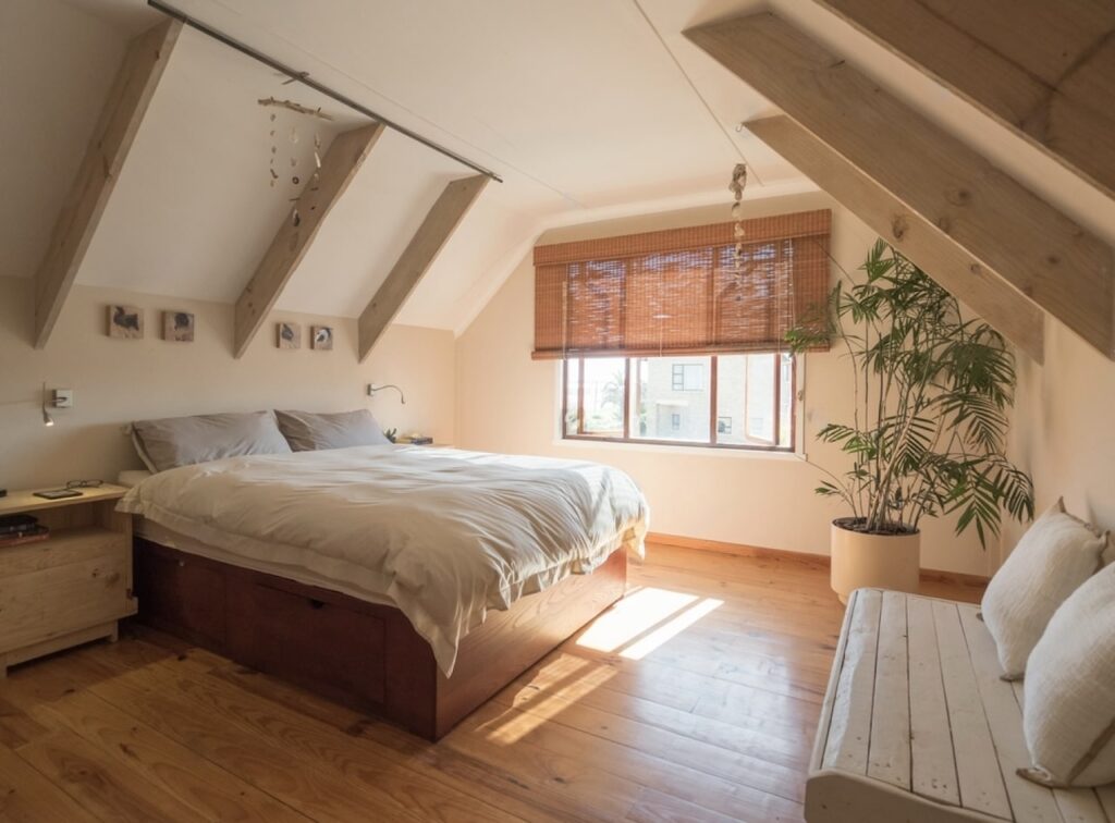 Cozy attic bedroom with natural light filtering through the window, featuring a large bed with beige bedding, exposed wooden beams, a bamboo window blind, and a potted plant adding a touch of greenery.