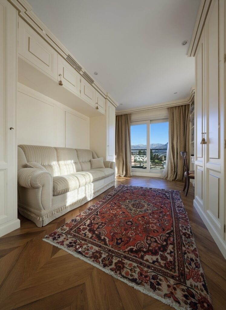 Elegant hallway leading to a balcony with a picturesque view, featuring classic design elements with a striped fabric sofa, ornate wall paneling, and a vibrant red traditional rug on herringbone hardwood flooring.