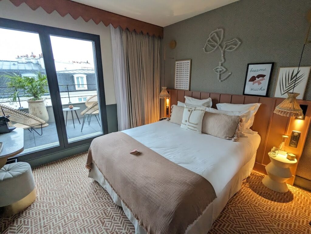 gay hotels in Paris: A chic and inviting hotel room featuring a large bed with white and beige linens, a decorative headboard, and a variety of pillows. Artistic wall decorations, including a line art face sculpture and framed prints with the text 'besame mucho,' complement the textured gray wall, while a balcony with outdoor seating offers a view of urban rooftops. The room's design is a harmonious blend of comfort and contemporary style.