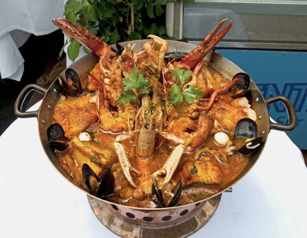 Marseille Food Guide: A lavish seafood platter featuring lobster, mussels, and various shellfish in a rich, herb-infused sauce, served in a large metal cooking pot, ready for a feast, garnished with fresh parsley on top.