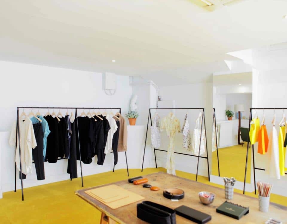 A bright and modern fashion studio, The Broken Arm in Paris, with clothes racks displaying a curated selection of chic apparel in neutral and vivid colors, alongside a creative workspace with design tools and sketches.