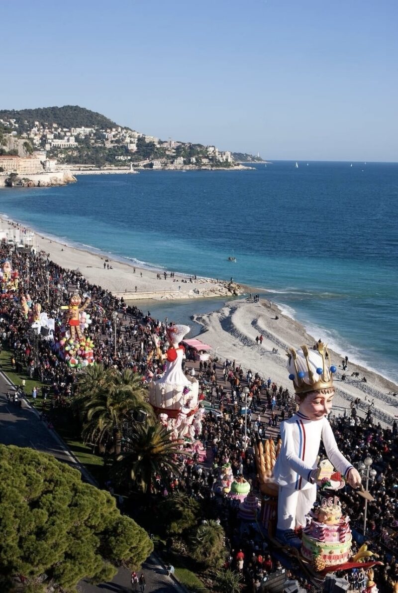 Aerial view of the Nice Carnival procession along the Promenade des Anglais, with whimsical floats including a chef figure and oversized desserts, throngs of people, and the azure Mediterranean Sea in the background.