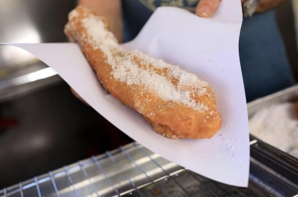 Marseille Food Guide: Close-up of a person's hands holding a freshly fried, sugar-dusted twist doughnut wrapped in white paper, with a blurred stainless steel counter in the background.