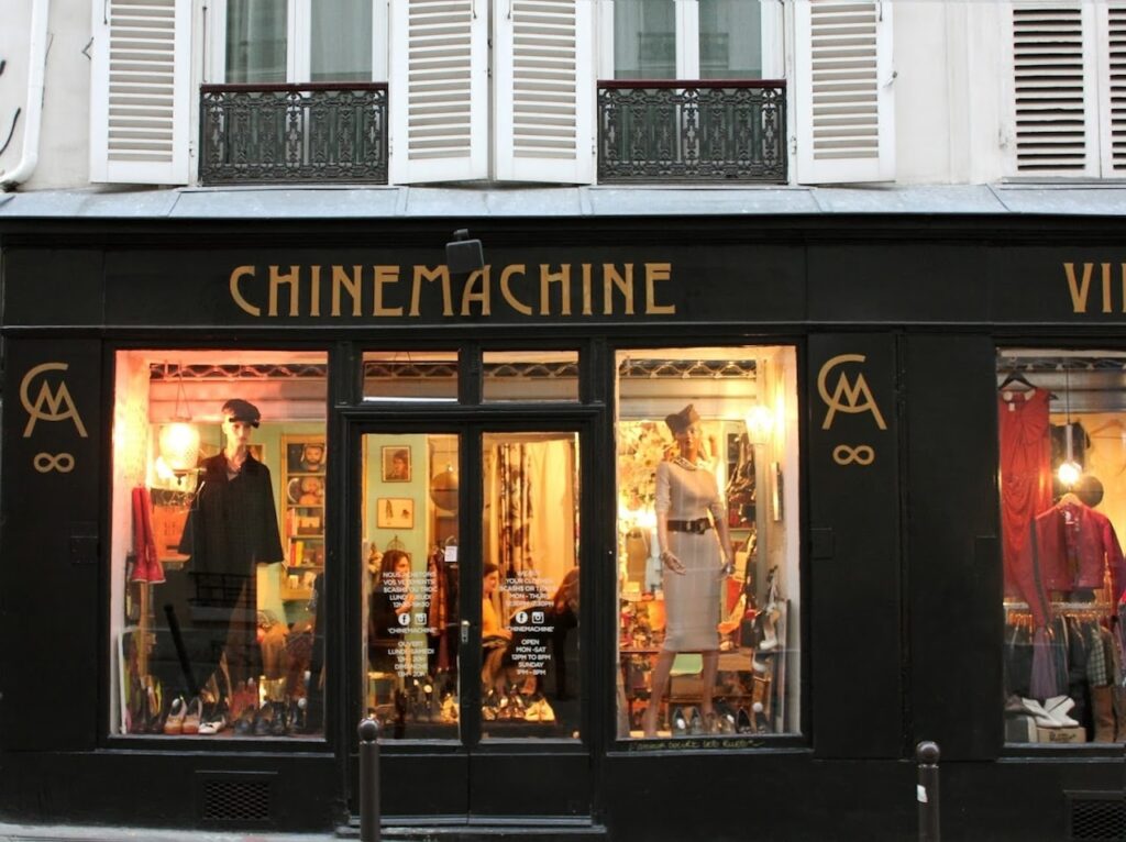 Vintage Shops in Paris: Exterior view of a vintage clothing store named 'CHINEMACHINE' in the evening, showcasing a variety of stylish garments and accessories in its warmly lit window displays against a classic Parisian building facade.
