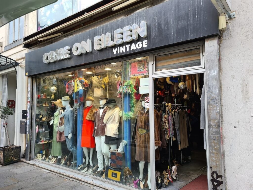 Street view of 'COME ON EILEEN VINTAGE' shop front with a display window full of eclectic vintage clothing and accessories, featuring mannequins dressed in colorful outfits from different eras, under a weathered shop sign.