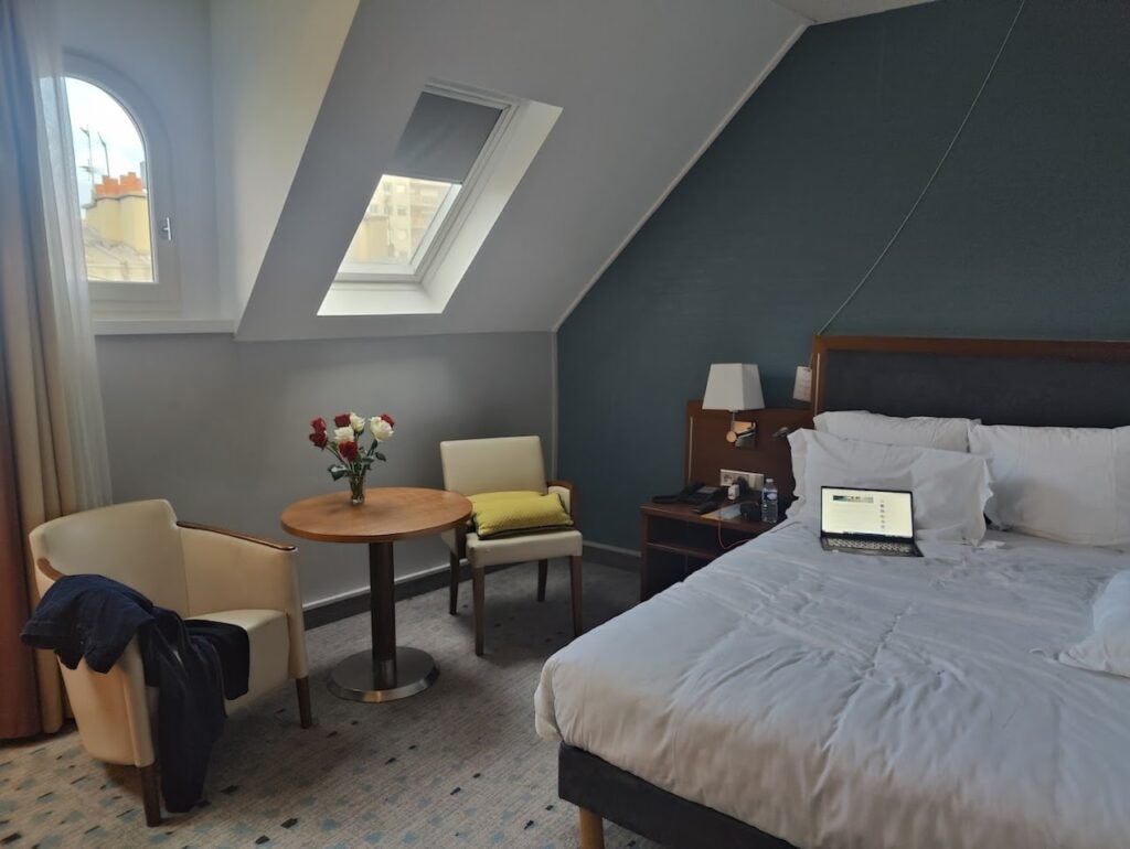 gay hotels in Paris: Attic-style hotel room with slanted ceiling and a skylight window, featuring a comfortable bed with a laptop open on it, a round table with two chairs and a vase of red and white roses, set against a calm grey and teal backdrop for a relaxed ambiance.