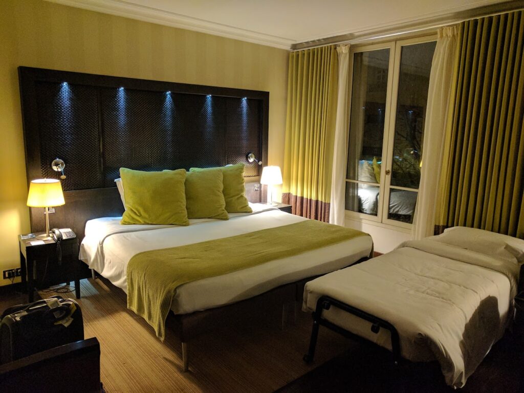 A cozy hotel room at night with a large double bed adorned with green accent pillows and a throw, a single bed to the right, and bedside lamps providing a warm glow. The room features a textured headboard and floor-to-ceiling windows with yellow drapes.