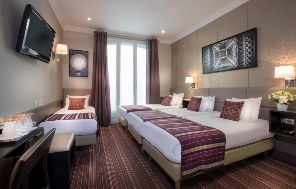 Bright and elegant twin room in a Paris hotel featuring two single beds with white linens and burgundy runners, a wall-mounted flat-screen TV, and decorative artwork. The room is completed with a work desk, plush curtains framing a balcony door, and tasteful ambient lighting.