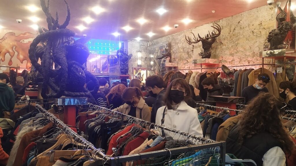 Interior of a bustling vintage clothing store, Free'P'Star, filled with shoppers browsing through racks of assorted clothing. An eye-catching large black sculpture of a mythical creature hangs from the ceiling, contributing to the shop's eclectic and artistic atmosphere.