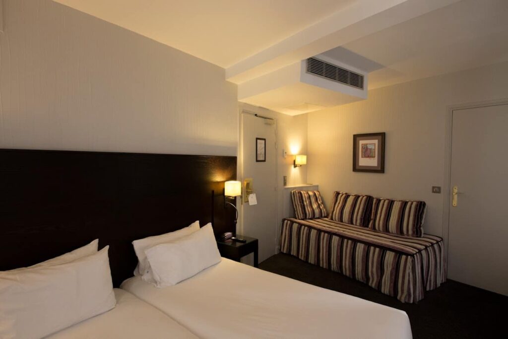 A neat and compact hotel room with twin beds, clean white bedding, and a dark headboard, complemented by a striped sofa bed. The room's decor includes soft wall-mounted lights, framed artwork, and an air conditioning unit, offering a cozy and functional space for travelers.