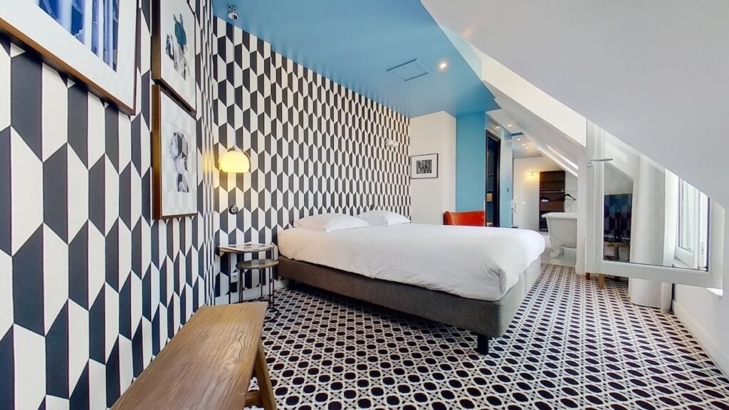 gay hotels in Paris: Eclectic hotel room with bold black and white geometric wallpaper, a simple bed with white linens, and a patterned floor. The slanted ceiling and blue accent wall add character, while framed art and a glimpse of an en-suite bathroom with a freestanding tub provide a unique charm.