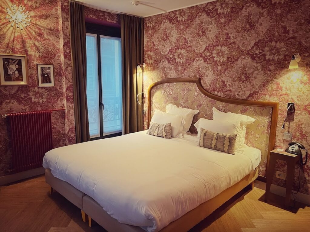 Vintage-inspired hotel room with an ornate red and gold damask wallpaper, matching drapery, and a classic wood-framed bed with plush white bedding. The room's warm lighting and traditional decor evoke a romantic Parisian ambiance.