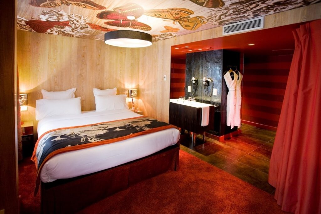 Vibrant hotel room with a whimsical butterfly ceiling mural, a bed with a bold orange throw, and wood-paneled walls. The open-plan design features a reflective bathroom area with a red-striped wall, creating a dynamic and visually stimulating space.