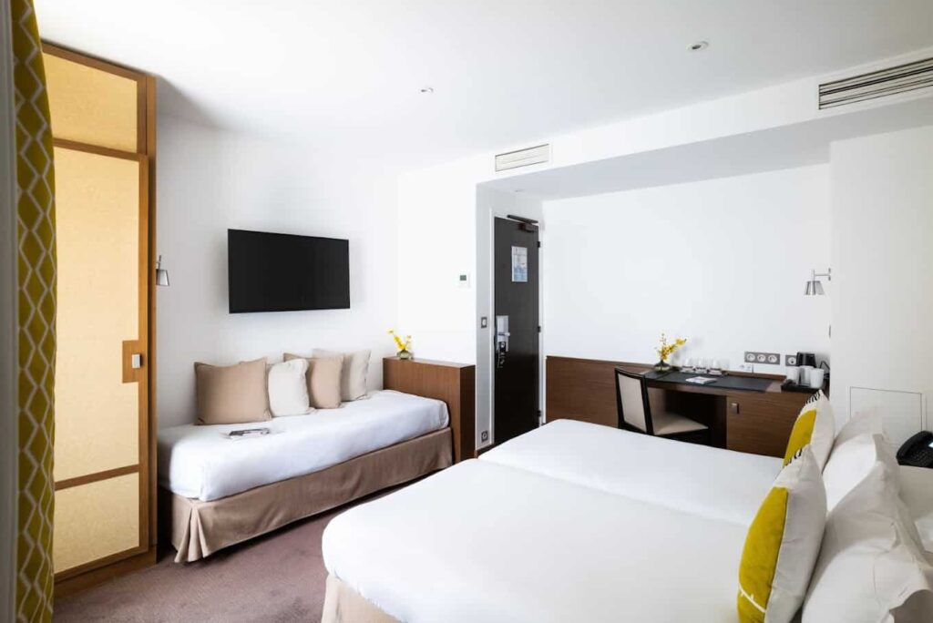 A bright, contemporary hotel room with a large bed and a single bed, both adorned with neutral tones and a pop of yellow from decorative pillows. The room features a sleek, wall-mounted television, a well-appointed work desk with a small flower arrangement, and an inviting, minimalist aesthetic.