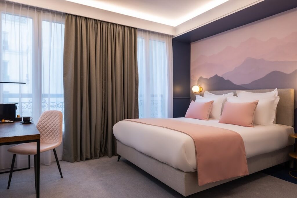 A cozy hotel room with a queen-sized bed with white and salmon pink bedding, a mountain mural on the wall, and elegant dark curtains framing a window. The room features a modern aesthetic with a plush chair and desk, creating a welcoming atmosphere for rest and relaxation.