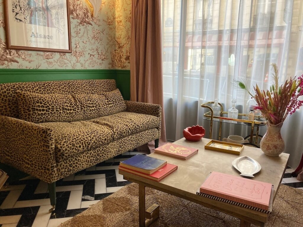gay hotels in Paris: Quirky lounge area with a leopard print sofa against a green trim, vintage pastoral wallpaper, and herringbone black and white flooring. The eclectic decor is accented with a marble coffee table adorned with books, a glass bar cart, and a vase of colorful flowers, contributing to a chic, Parisian vibe.