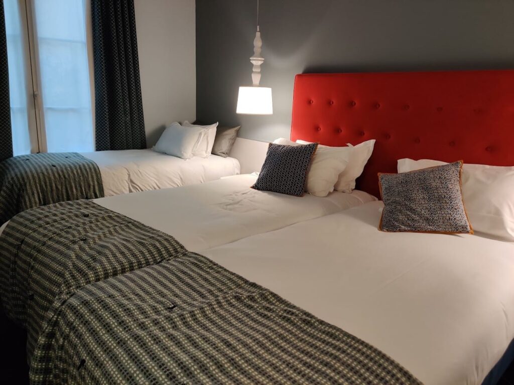 A chic hotel room with a bold red tufted headboard contrasting against crisp white bedding. The beds are accented with houndstooth bedspreads and eclectic throw pillows, flanked by dark patterned curtains and soft lighting, creating an atmosphere of modern elegance with a classic touch.