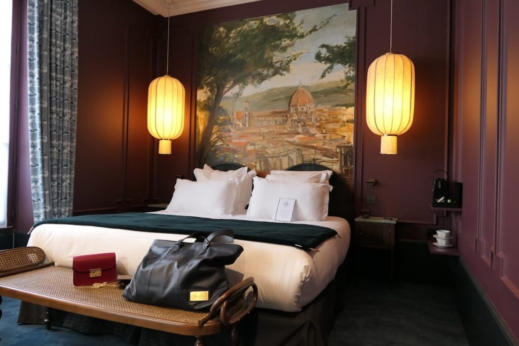 Luxurious hotel room with rich aubergine walls, a plush bed with white linens, and an oversized painting of a Tuscan landscape as the headboard. The room is accessorized with a leather bench holding a travel bag and a stylish box, flanked by warm hanging lanterns, enhancing the room's opulent and travel-inspired theme.