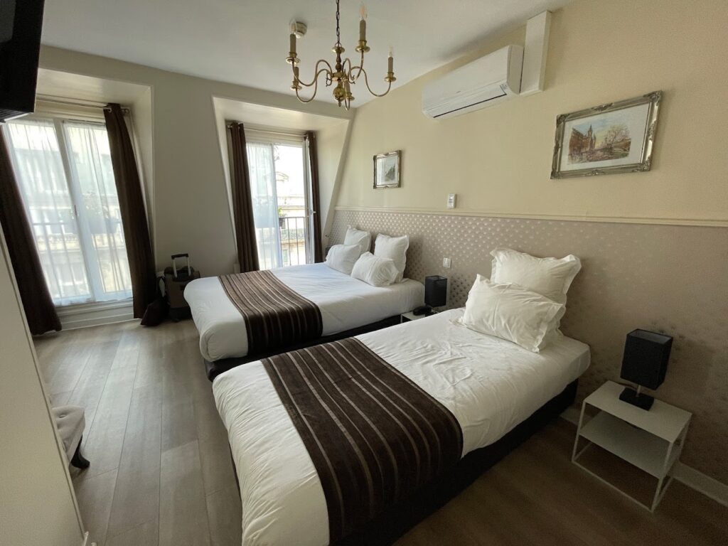 A well-lit, comfortable twin room with an airy ambiance, featuring two beds with white linens and chocolate brown accents. The room includes a classic chandelier, air conditioning units, framed artwork on the walls, and balcony doors allowing natural light to enhance the serene, inviting space.