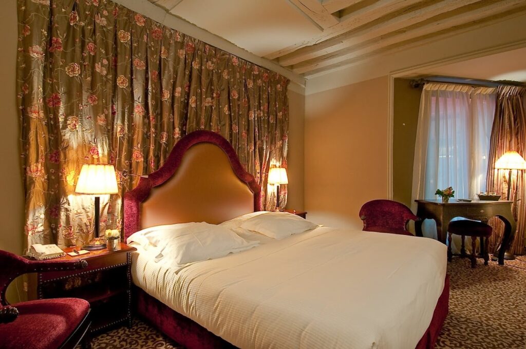 A romantic and vintage-styled hotel room featuring a large bed with a burgundy velvet headboard and matching drapes with a floral pattern. Exposed wooden ceiling beams add character to the room, which also includes classic furniture, a desk with a lamp, and sheer curtains that allow for a warm, diffused light.