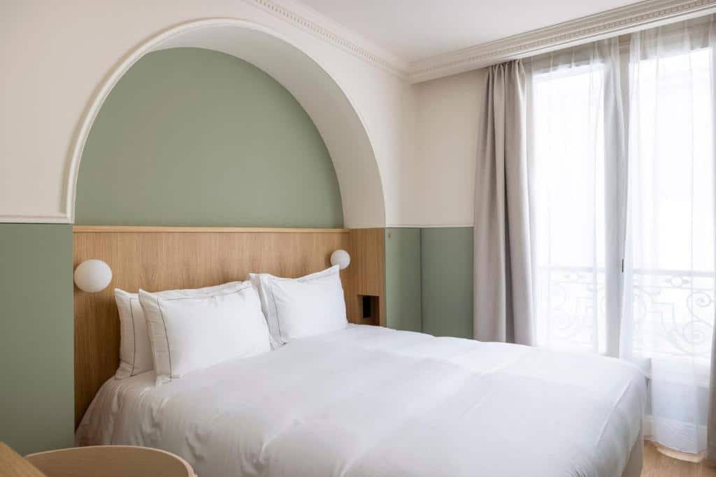 gay hotels in Paris: Minimalist and serene hotel room with a curved sage green wall over a light wood headboard, crisp white bedding, and spherical bedside lamps. The room's high ceiling and flowing white curtains give it an airy and open feel, complementing the clean, modern design.