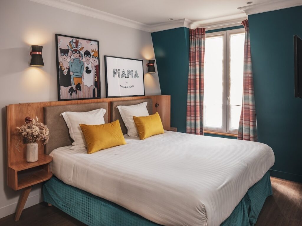 Modern hotel room with a deep teal bed frame, contrasting mustard yellow pillows, and mid-century modern wood furnishings. The playful and artistic vibe is emphasized by whimsical framed artwork above the bed and tartan curtains that add a touch of tradition to the space.