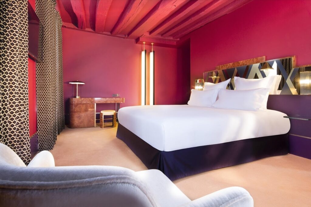 A luxurious hotel room with a vibrant fuchsia wall and exposed red ceiling beams, creating a bold statement. The room features a large bed with crisp white linens, an artistic geometric headboard, and a unique floor lamp with vertical lights. Completing the opulent decor are a plush velvet chair, animal print drapes, and a vintage-inspired desk, blending contemporary design with classic glamour.