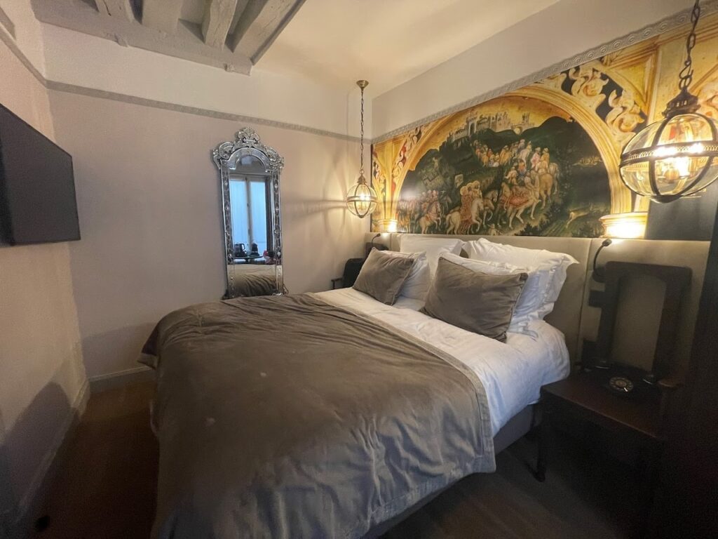 gay hotels in Paris: Elegant hotel room with a classic fresco mural above the bed, ornate hanging lanterns, and a tall baroque-style mirror reflecting the room's charm. The bed is dressed in luxurious grey bedding, and the exposed wooden beam adds a rustic touch to the sophisticated decor.