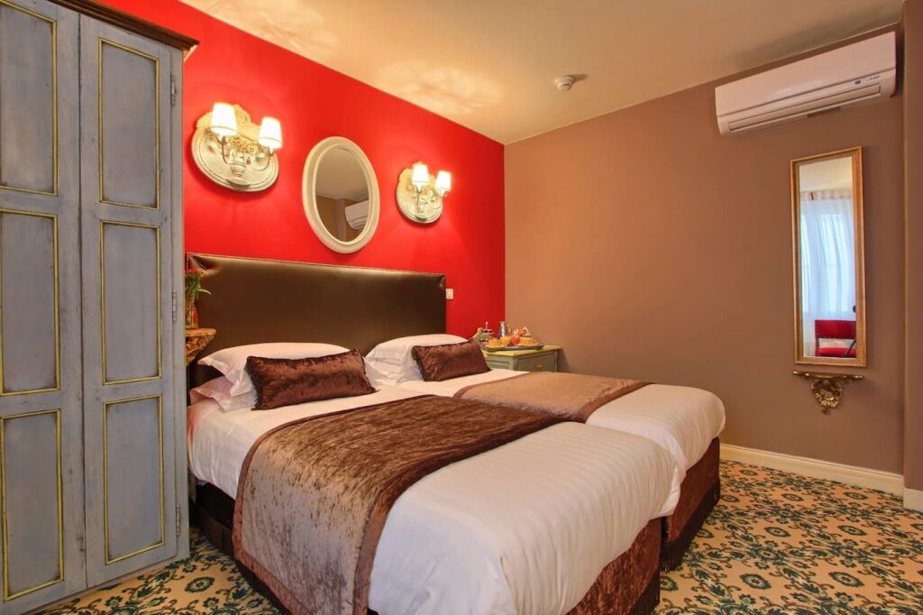 Vibrant hotel room with a striking red accent wall and two single beds with plush brown runners and pillows. Vintage style is reflected in the distressed wardrobe, ornate gold-framed mirrors, and classic patterned carpet, alongside a tray with a teapot and cups hinting at a cozy atmosphere.